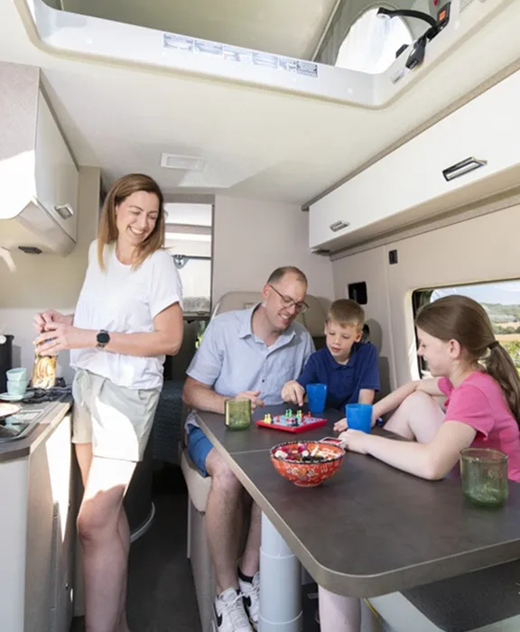 Familie im Campingbus: Vater und Kinder spielen am Tisch, Mutter lächelt in der Küche.