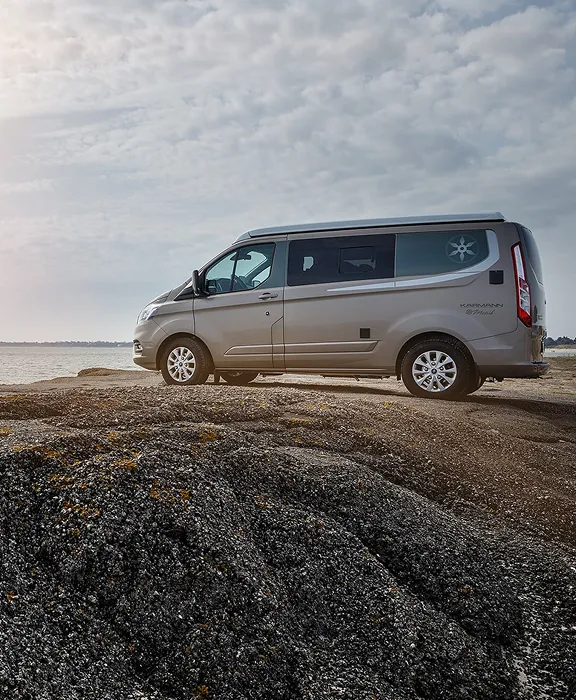 Karmann-Mobil Campingbus steht auf felsigem Küstenabschnitt mit Blick auf das Meer unter bewölktem Himmel und leichtem Sonnenlicht.